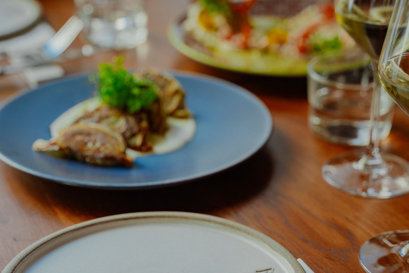 A dinner course on a blue plate beside glasses of white wine, the next plate waiting