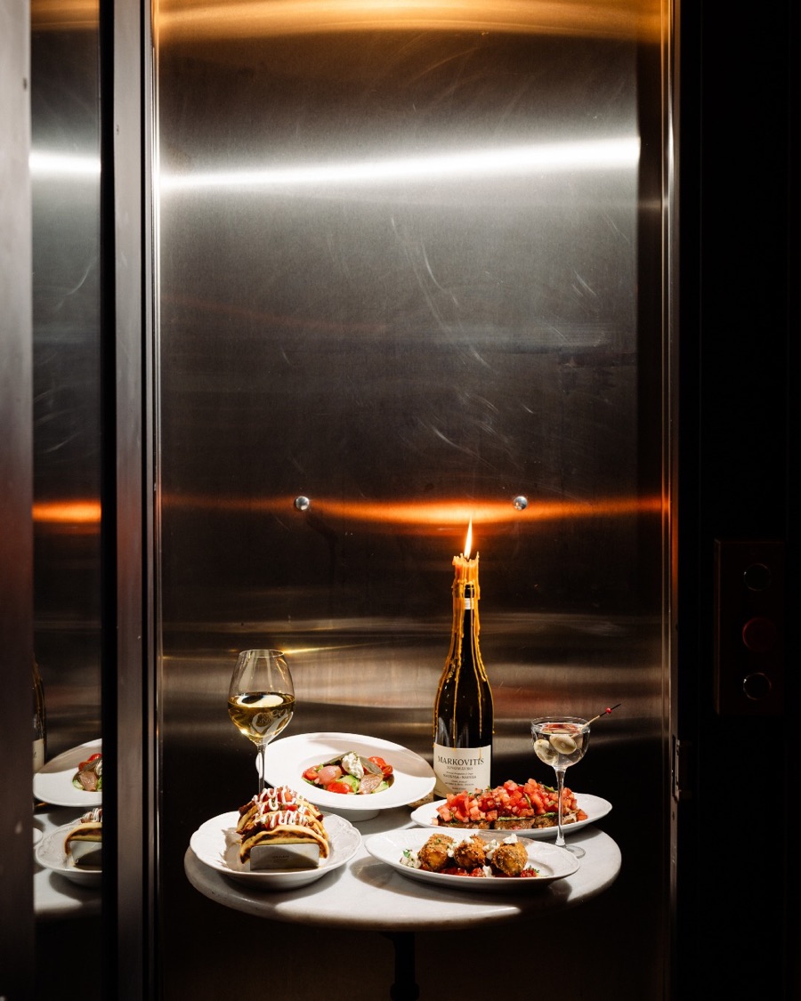A candlelit table at Eleve wine bar: a bottle, a glass of white and small plates against a steel wall